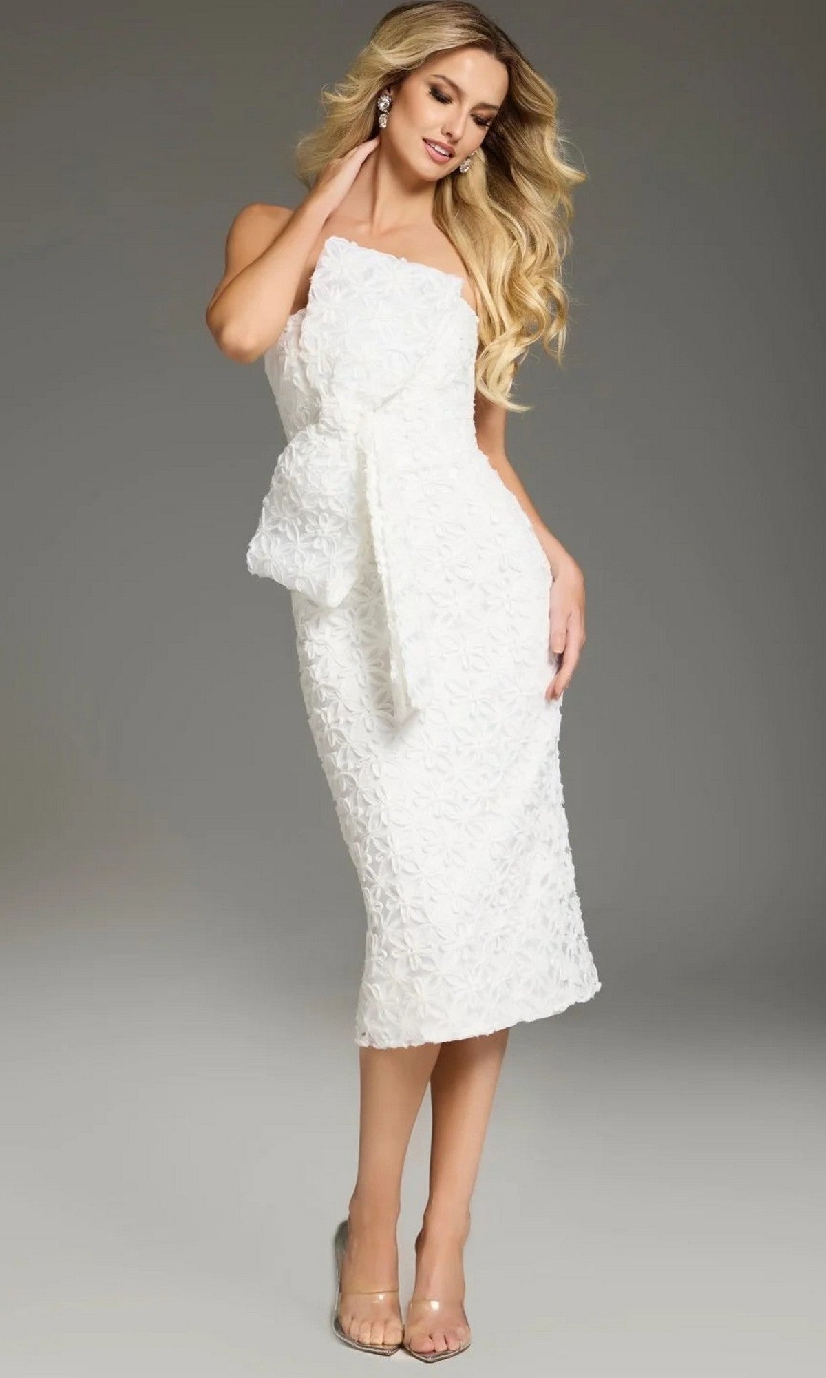 A woman stands against a plain gray background wearing the Jovani 39076 Midi Wedding Guest Dress, a textured white strapless formal dress with a large side bow, styled with clear high heels.